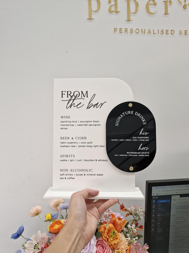 May include: A white arched acrylic sign with black text titled FROM the bar, listing wine, beer, cider, spirits, and non-alcoholic options. Attached is a black oval acrylic sign titled SIGNATURE DRINKS with his and hers cocktail recipes.