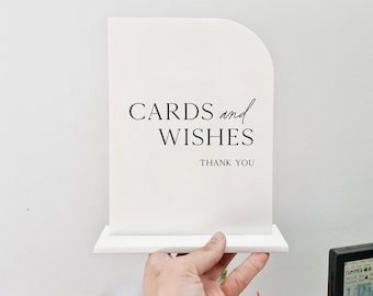 May include: A white, arched sign with the words "CARDS and WISHES THANK YOU" in black lettering. The sign is held in front of a white wall, with a bouquet of colorful flowers at the bottom.