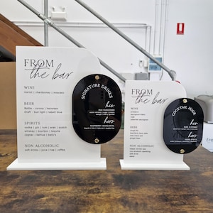 May include: Two white acrylic bar menu signs with rounded corners, featuring black text and attached black oval acrylic plaques. The signs list drink categories including wine, beer, spirits, and non-alcoholic options, with "From the bar" and "Signature Drinks" or "Cocktail Hour" headings.