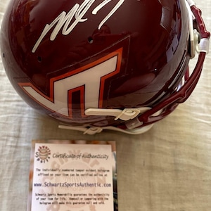 May include: A glossy maroon football helmet featuring a white and orange VT logo on the side and a silver autograph on the top. Beside the helmet is a rectangular white Certificate of Authenticity card from Schwartz Sports Memorabilia.