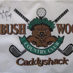 May include: A white golf flag featuring the Bushwood Country Club logo with a brown gopher, crossed golf clubs, and a golf ball. The text reads Bushwood Country Club and Caddyshack, with a handwritten signature in the upper left corner.