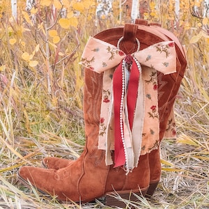 May include: A pair of brown suede cowboy boots featuring decorative stitching on the shaft and toe. A large bow made of floral-patterned fabric, solid red ribbon, and a string of white beads is attached to the top of the boot with a metal ring.