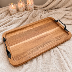 Puede incluir: Una bandeja de servicio rectangular de madera con un acabado de grano de madera marrón claro natural y dos asas de metal negro con un diseño trenzado. La bandeja tiene esquinas redondeadas y una superficie plana y lisa para objetos.