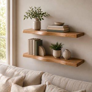 May include: Two horizontal wall-mounted shelves made of natural light brown wood with live edges. The top shelf holds a ceramic vase with greenery and a stack of three books topped by a bowl. The bottom shelf holds five books, a stone bookend, a potted plant, and a ribbed ceramic vase.