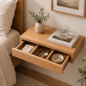 May include: A light oak floating nightstand with a single drawer divided into three compartments. The drawer contains a small fabric pouch, black-rimmed glasses, a wristwatch, and a ceramic bowl with jewelry. A vase, a book, and a glass sit on top.