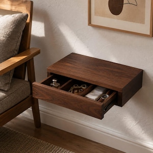 May include: A dark brown wooden floating wall shelf featuring a single pull-out drawer divided into three compartments. The drawer contains a small glass bottle, a gold chain necklace, a gold ring, a folded white cloth, and a wristwatch.