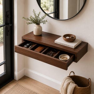 May include: A dark wood floating wall shelf with a single pull-out drawer divided into four compartments. The shelf holds a beige ceramic vase with greenery, a book titled Live Beautifully, and a small wooden bowl containing a wristwatch.
