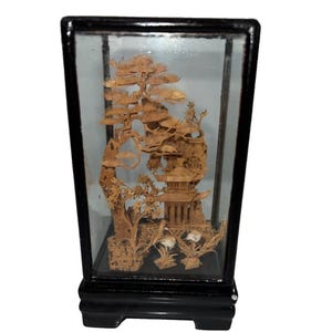 May include: A rectangular glass display case with a black frame containing a detailed cork carving. The carving depicts a multi-tiered pagoda, a large pine tree, and small bird figures set against a textured landscape of rocks and foliage.