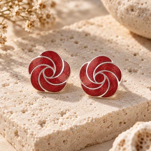 Può includere: Un paio di orecchini a lobo a forma di fiore stilizzato con un centro testurizzato rosso intenso e contorni in metallo color argento liscio. Il design è composto da segmenti vorticosi a forma di petalo che creano un aspetto circolare.