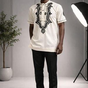 May include: A short-sleeved cream-colored shirt featuring a mandarin collar and intricate black embroidery around the neckline and sleeve cuffs, paired with solid black trousers and black dress shoes for a formal ensemble.