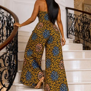 May include: A strapless jumpsuit featuring a mustard yellow base with a dark brown leaf-like pattern and circular floral motifs in shades of blue and peach. The garment has a fitted bodice and wide-leg trousers that fall to the floor.