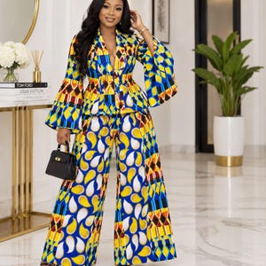 May include: A two-piece outfit consisting of a long-sleeved blazer with a deep V-neck and wide-leg trousers. The set features a vibrant geometric and paisley pattern in shades of blue, yellow, white, and red. A small black handbag is held in one hand.