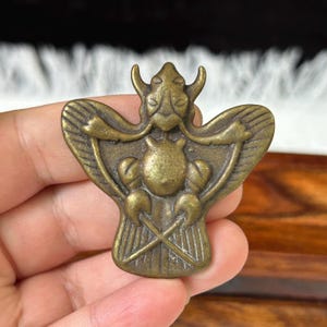 May include: A brass-toned metal ornament shaped like a stylised insect with prominent wings, a central rounded body, and two antennae. The surface features engraved lines on the wings and a cross-hatched pattern at the base of the figure.
