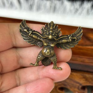May include: A small, antique-style brass figurine depicting a mythical creature with wings spread wide, a prominent headdress, and textured legs. The figure has a rounded torso and holds a snake-like form in each hand, finished in a dark patina.