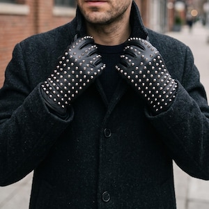 Puede incluir: Guantes de cuero negro con un patrón denso de pequeños remaches cónicos de metal plateado en el dorso de las manos y los dedos. Los guantes se llevan con un abrigo de lana texturizada gris carbón y un suéter negro.