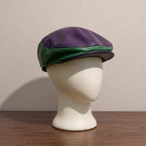May include: A flat cap constructed from panels of purple and green leather. The cap features a rounded crown and a short, stiff brim at the front. The leather has a visible grain texture, and the two colors are joined by visible stitching.