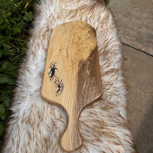 May include: A wooden serving board with a handle, featuring a natural light and dark brown wood grain pattern. The surface includes two distinct dark blue, jagged, lightning-like resin inlays that create a striking contrast against the wood.