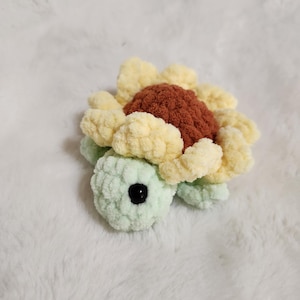 May include: A crochet turtle plushie featuring a light green body with a dark brown center and pale yellow petal-like shell. The turtle has a rounded head with a single visible black plastic eye and a soft, textured yarn construction throughout.