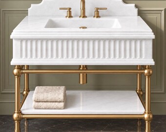 May include: A white marble bathroom vanity featuring a fluted front apron, a scalloped backsplash, and a gold-toned metal frame. The unit includes a rectangular sink, gold faucet hardware, and a lower marble shelf holding two folded beige towels.