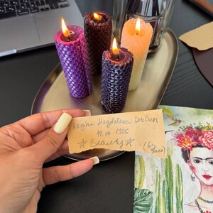 A person's hand holding a handwritten note, with a laptop, candles, and a framed image of a woman's face on a table in the background.