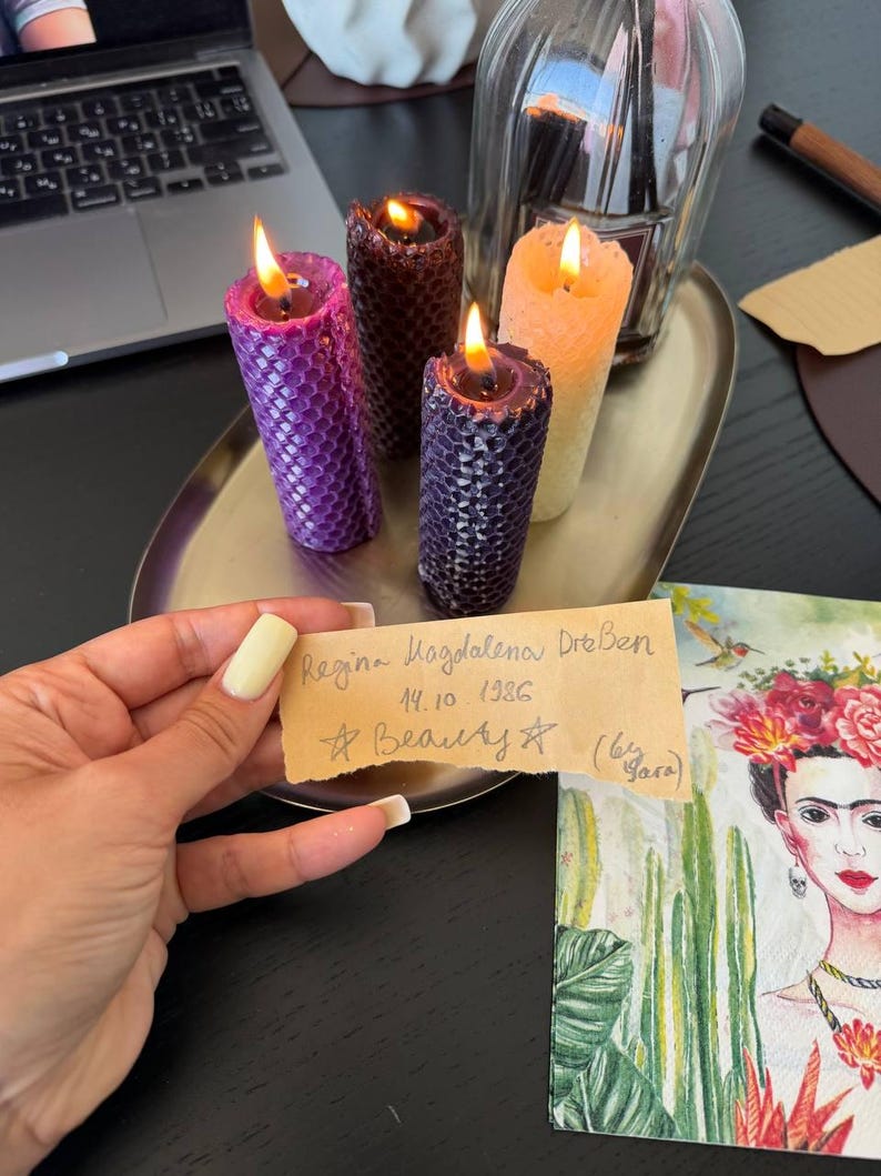 A person's hand holding a handwritten note, with a laptop, candles, and a framed image of a woman's face on a table in the background.