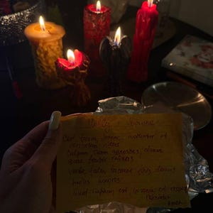 A hand holding a handwritten note in front of a collection of lit candles on a table.