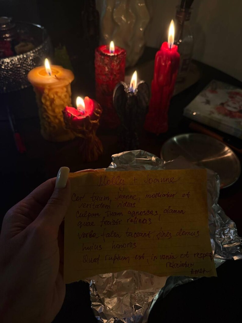 A hand holding a handwritten note in front of a collection of lit candles on a table.