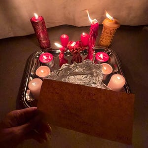 A hand holds a handwritten note in front of a tray with lit candles, creating a warm and intimate atmosphere.