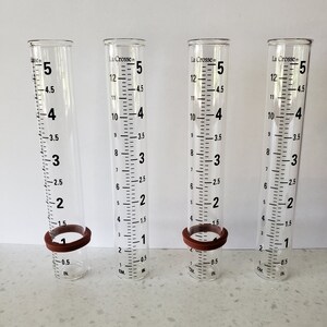 May include: Four clear glass rain gauges with black markings and brown rubber rings. The gauges measure in inches and centimeters. La Crosse is printed on the gauges.