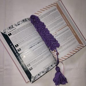 Peut inclure: Un marque-page en crochet violet avec un motif texturé en forme de feuille et un pompon en fil assorti à l'extrémité, posé sur un livre ouvert dont les pages visibles portent les titres A Woman of Character et Overcoming Obstacles.