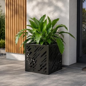 Pode incluir: Um vaso quadrado de metal preto com um padrão decorativo de ripas diagonais cortadas a laser. O vaso contém uma planta lírio-da-paz verde e exuberante com folhas largas e pontiagudas, criando um contraste com o recipiente geométrico escuro.