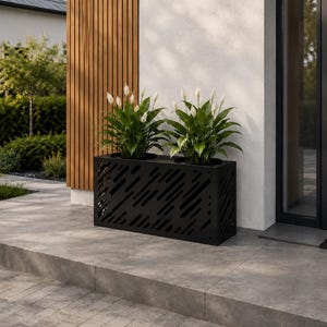Op de afbeelding: Een rechthoekige zwarte metalen plantenbak met een decoratief diagonaal lattenpatroon. De bak bevat twee groene lepelplanten met witte bloemen en staat op een grijs betegeld terras bij een moderne ingang van een gebouw.