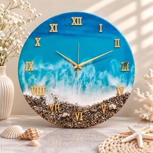 May include: A round wall clock featuring a blue and white ocean wave design with gold Roman numerals and gold hands. The bottom section of the clock face is textured with brown sand and small white seashells, creating a beach-inspired aesthetic.