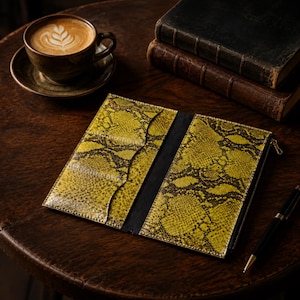 May include: A long, rectangular wallet featuring a vibrant yellow and black snakeskin pattern. The interior displays multiple card slots on the left side and a zippered compartment on the right, with a black central spine and a gold-toned zipper pull.