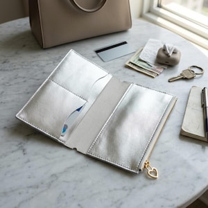 May include: A metallic silver wallet with an open interior featuring multiple card slots and a zippered compartment. The zipper pull is shaped like a small gold heart. The wallet is laid flat, revealing its reflective surface and stitching.