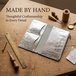 May include: A silver-colored wallet with a textured crocodile-skin pattern, featuring multiple interior card slots and a zipper closure. The text "MADE BY HAND" and "Thoughtful Craftsmanship in Every Detail" is visible above the wallet.