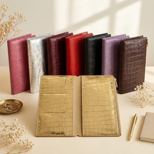 Puede incluir: Una colección de carteras rectangulares con textura de cocodrilo, presentadas en varios colores incluyendo rosa, plata, burdeos, rojo, negro, morado y marrón, con una cartera dorada abierta en primer plano mostrando ranuras para tarjetas.