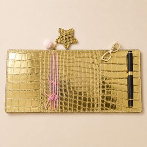 May include: A gold metallic faux leather notebook cover with a crocodile skin texture. It features three vertical pink elastic bands, a gold star charm with a small pink pom-pom, a gold-toned glasses-shaped clip, and a black pen with gold trim.