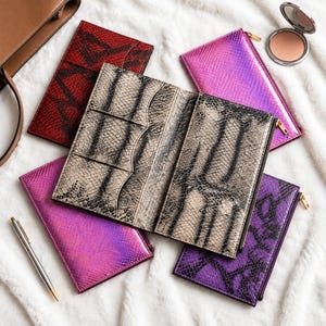 Pode incluir: Uma coleção de carteiras e bolsas retangulares com várias texturas e cores, incluindo uma carteira central aberta com padrão de pele de cobra bege e preta, uma bolsa vermelha, duas bolsas rosa iridescentes e uma bolsa roxa.