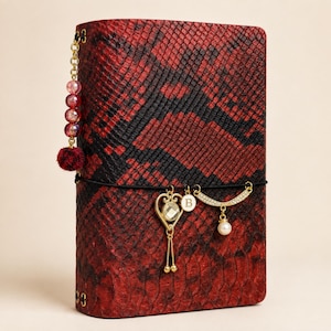 May include: A rectangular notebook with a red and black snakeskin pattern cover. A gold chain with four beads hangs from the top left. A black elastic band wraps around the center, holding three gold charms: a heart with a crystal, a letter B, and a crescent moon with a pearl.