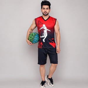 Op de afbeelding: Een mouwloos shirt met V-hals in een rood-naar-zwart verloop met een witte afbeelding van een basketballer in beweging, gecombineerd met een effen zwarte sportshort en een groen met blauw gemanipuleerde basketbal in de hand.