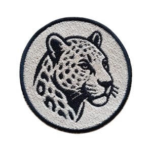 May include: A circular embroidered patch featuring a black and white leopard head design. The leopard is depicted in profile facing right, with detailed black spots against a light gray background, all enclosed within a thick black border.