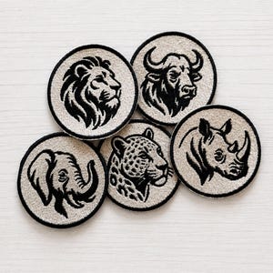 Op de afbeelding: Vijf ronde geborduurde patches met zwarte draadillustraties van een leeuw, een buffel, een olifant, een luipaard en een neushoorn op een lichtbeige stoffen achtergrond. Elke patch heeft een dikke zwarte gestikte rand.
