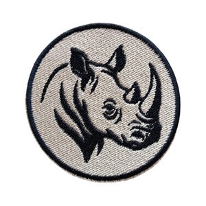 May include: A circular embroidered patch featuring a black line art illustration of a rhinoceros head on a light beige background. The design is enclosed within a thick, dark black stitched border, creating a clean and defined circular shape.
