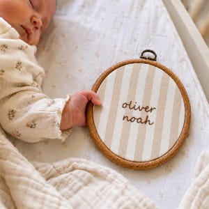 Könnte beinhalten: Ein runder Stickrahmen aus Holz mit einem Stoff mit vertikalen beigen und weißen Streifen. Der Name oliver noah ist in braunem Garn in die Mitte des Stoffes gestickt. Der Rahmen hat oben eine kleine Metallschraube zum Festziehen.
