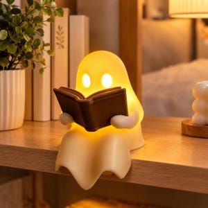 May include: A glowing, cream-colored ghost-shaped lamp made of smooth material, featuring two oval-shaped eyes and holding a small, dark brown open book in its hands. The lamp emits a warm, soft light from within its rounded, translucent body.