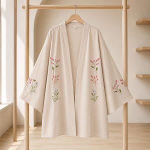 May include: A beige open-front kimono jacket featuring long, wide sleeves and embroidered floral motifs. The embroidery displays pink flower buds, green stems, and white daisies arranged vertically along the front panels and on the sleeves.