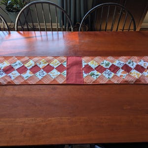 May include: A rectangular table runner featuring a patchwork design of diamond-shaped fabric pieces in shades of orange, rust, and white. The pattern includes floral and plaid prints, with a solid rust-colored center panel and orange trim.