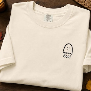 May include: An ivory-colored short-sleeve t-shirt featuring a small black graphic of a ghost on the left chest area. The ghost has a simple line-drawn face and the word "Boo!" printed underneath it in a black, handwritten-style font.