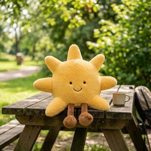 May include: A plush sun toy with a bright yellow, textured surface, featuring a simple embroidered face with two small black eyes and a curved mouth, along with two dangling brown legs with rounded feet, sitting on a wooden surface.